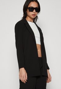 Fekete zakó, amelyek testreszabott szabású, sima anyagból készült. Hosszú ujjakkal és kerek gallérral rendelkezik. Fehér crop top fölött viselik.