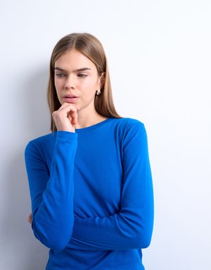 Nuori nainen sinisessä pitkähihaisessa paidassa, käsi leualla, katsomassa mietteliäästi sivulle valkoista taustaa vasten.