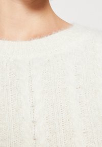 Großer Strickpullover in Off-White mit weitem Rundhalsausschnitt, weichem Material und gerippten Bündchen, der ein vertikales Muster zeigt.