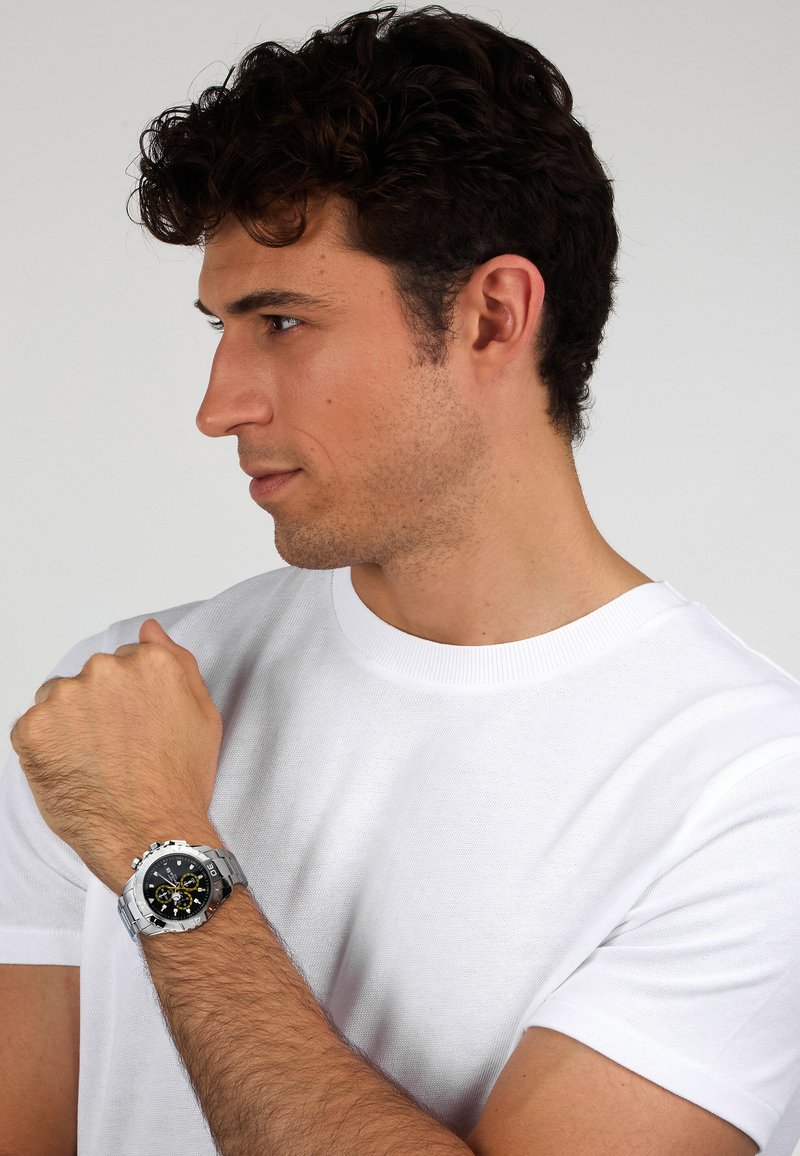 Orologio cronografo argento con quadrante nero, dettagli gialli e bracciale in metallo, indossato su un polso contro uno sfondo di t-shirt bianca.
