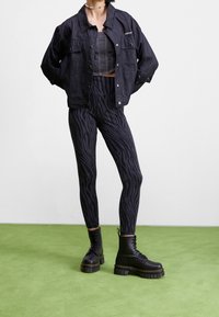 Svart denimjacka över en åtsittande svart crop top, ihop med svarta leggings med zebramönster och kraftiga svarta stövlar. Grönt golv.