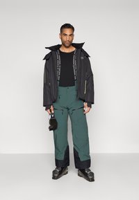 Casaco de esqui preto com detalhes amarelos, camisa de manga longa preta, calças de esqui verdes com punhos azul-escuro e botas de esqui pretas. A segurar pequenos óculos de esqui.