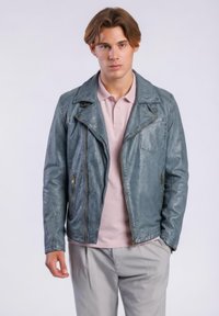 Junger Mann, der eine blau-graue Lederjacke über einem hellrosa Poloshirt und hellgrauen Hosen vor einem einfachen weißen Hintergrund trägt.