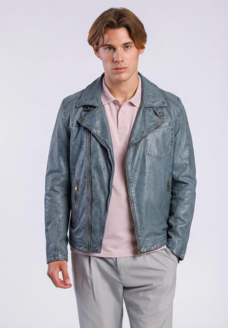 Junger Mann, der eine blau-graue Lederjacke über einem hellrosa Poloshirt und hellgrauen Hosen vor einem einfachen weißen Hintergrund trägt.