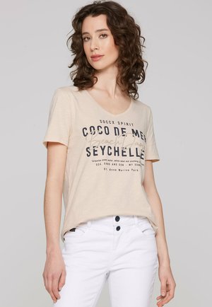 Beige T-Shirt mit kurzen Ärmeln und V-Ausschnitt, versehen mit dunkelblauen Textgrafiken. Kombiniert mit weißen Hosen, die einen lässigen Schnitt zeigen.