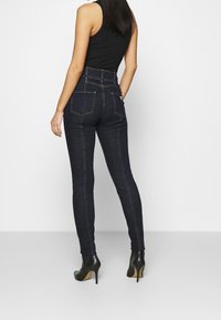 Jeans de denim oscuro de cintura alta con un corte ajustado, cinco bolsillos y costuras en contraste, combinados con botas de tacón negro hasta el tobillo.