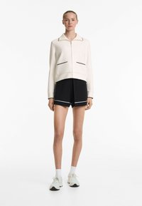 Light cream zip-up jacket with black piping, long sleeves, and two front pockets, paired with black shorts featuring white accents and sneakers.