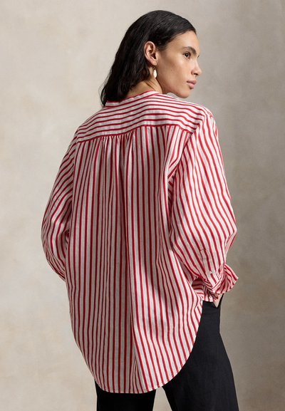 Žena s tmavými vlasmi, oblečená v červeno-bielej blúzke s vertikálnymi pruhmi s dlhým rukávom a čiernych nohaviciach, pózujúca s rukami v vreckách pred neutrálnym pozadím.