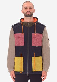 Uomo che indossa una giacca multicolore con zip, con maniche beige, corpo blu navy, tasche superiori rosse, tasche inferiori gialle e lacci arancioni.