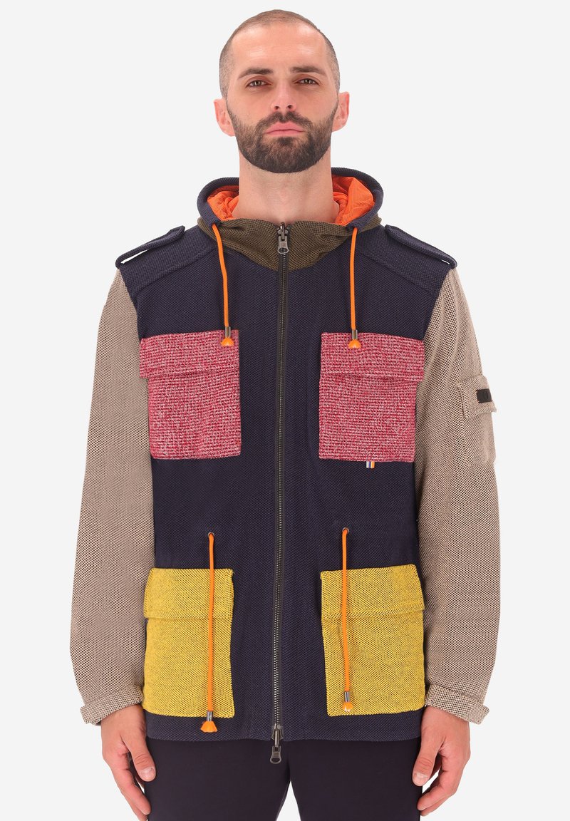 Uomo che indossa una giacca multicolore con zip, con maniche beige, corpo blu navy, tasche superiori rosse, tasche inferiori gialle e lacci arancioni.