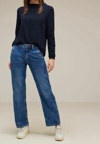 Tmavě modrý top s dlouhým rukávem v kombinaci s modrými džíny se širokými nohavicemi, které mají klasickou denimovou texturu a zapínání na knoflíky, doplněné bílými teniskami.