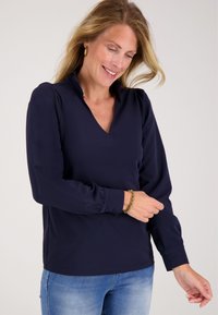 Vrouw die glimlacht en haar marineblauwe blouse met lange mouwen en V-hals recht trekt, met gouden oorbellen en armband, gecombineerd met lichtblauwe spijkerbroek.