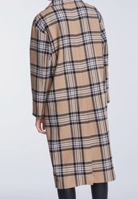 Manteau en laine à carreaux beige avec des accents noirs et blancs, doté d'une coupe décontractée, de manches longues et d'une fente au dos pour faciliter les mouvements.