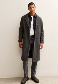 Grauer Wollmantel mit Reverskragen, getragen über einem weißen T-Shirt und einem marineblauen Hemd mit Knöpfen, kombiniert mit grauen Jeans und braunen Stiefeln.