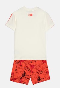 T-shirt bianca in cotone con accento rosso sulla manica; abbinata a pantaloni corti rossi con stampe di personaggi Disney neri, tra cui Topolino.