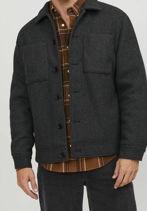 Man draagt een donkergrijs wollen jasje over een bruin geruit overhemd met knopen en zwarte broek, staat met ontspannen handen.