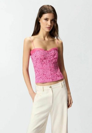 Donna che indossa un top di pizzo rosa con spalline sottili e pantaloni crema a vita alta, in piedi con una mano in tasca.