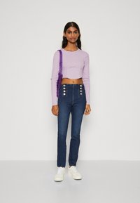 Suéter recortado de color púrpura claro con mangas largas, combinado con jeans de cintura alta en azul oscuro que presentan acentos de botones plateados en la parte delantera. Zapatillas blancas.