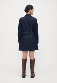 Σκούρο μπλε τζιν φόρεμα με εφαρμοστή σχεδίαση, κλείσιμο με κουμπιά και ελαφριά ραφή. Συνδυάζεται με ψηλές καφέ δερμάτινες μπότες που έχουν αγκράφες.