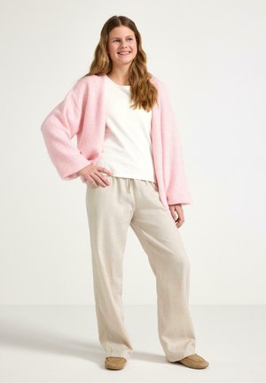 Teini-ikäinen tyttö hymyilee, yllä vaaleanpunainen neuletakki valkoisen paidan päällä, beigejä leveälahkeisia housuja ja ruskeat liukusandaalit, seisoo käsivarressaan.