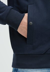 Sudadera azul marina con puños y dobladillo acanalados. Bolsillo lateral con un detalle de botón negro. Textura de tela suave con un diseño minimalista.