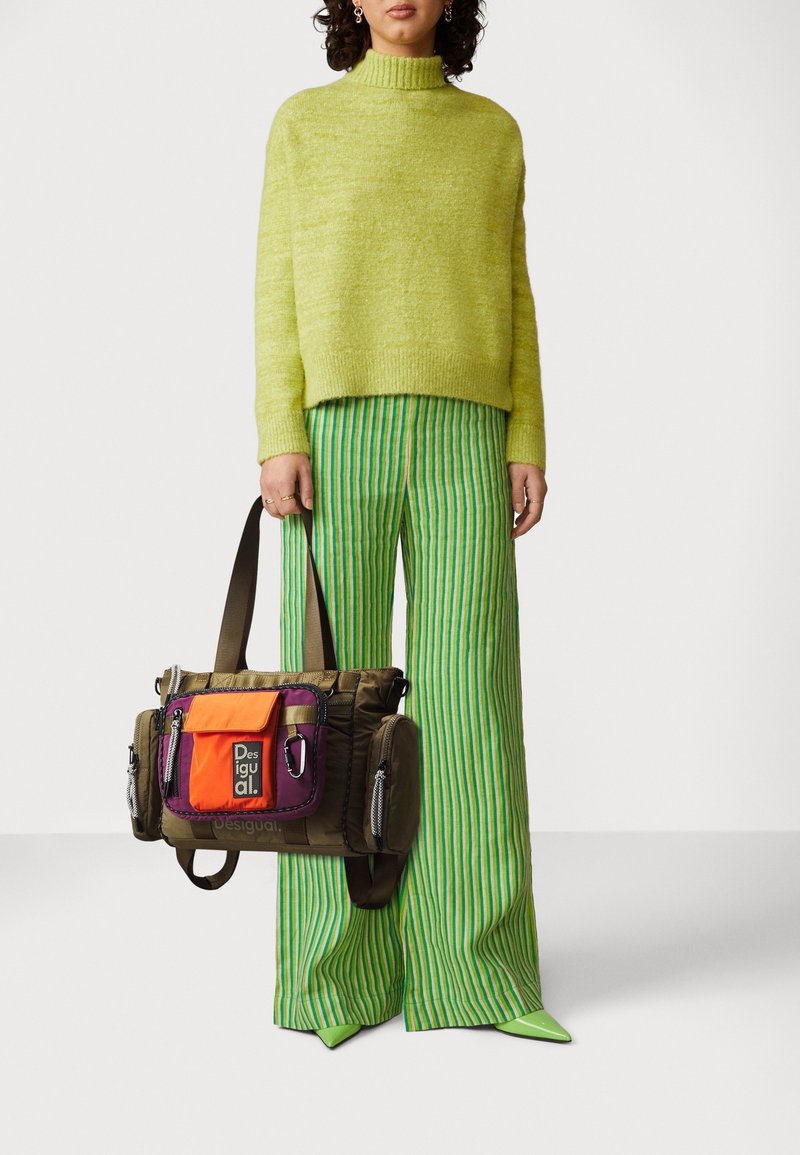 Personne portant un col roulé duveteux vert clair, un pantalon large à rayures verticales vertes et bleues, tenant un sac multicolore vert, orange et violet.