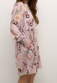 Vestido floral en rosa con mangas largas, puños fruncidos y bolsillos laterales. Presenta una mezcla de patrones en púrpura, amarillo y verde sobre un fondo texturizado.