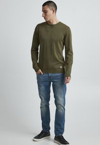 Olivno zeleni pulover dolgi rokavi z rebrastim ovratnikom in manšetami, v kombinaciji s svetloblujnimi jeans hlačami in črnimi športnimi čevlji, stoji na nevtralni podlagi.
