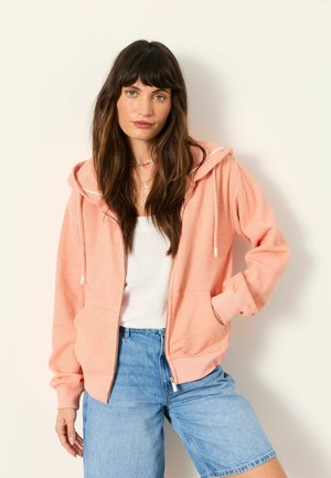 Žena s dlhými tmavými vlasmi, ktorá má na sebe broskyňovú mikinu na zips, biely top a riflové kraťasy, stojaca pred jednoliatym svetlým pozadím.