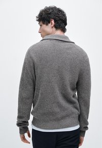 Maglione marrone lavorato a maglia con ampio colletto e polsini a coste, caratterizzato da un motivo a trama e una vestibilità comoda. Indossato sopra una camicia bianca.