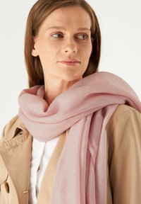 Écharpe rose clair en tissu doux et lisse, drapée gracieusement autour du cou, avec un léger éclat. Portée sur une veste beige.