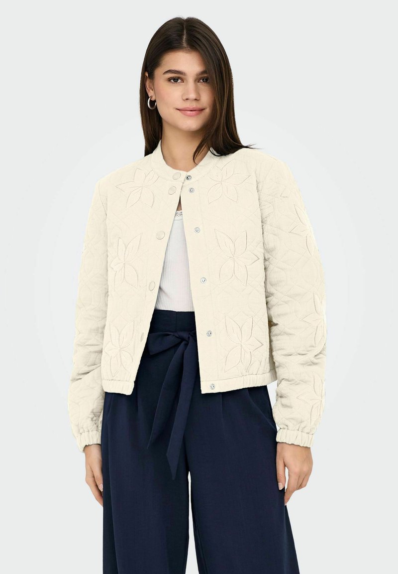 Jeune femme portant une veste matelassée crème à motifs floraux, un haut blanc et un pantalon taille haute bleu marine avec une ceinture nouée.