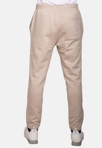 Pantaloni sportivi beige con cintura elastica, tasche laterali e una tasca sul retro, abbinati a scarpe da ginnastica bianche. Tessuto liscio.