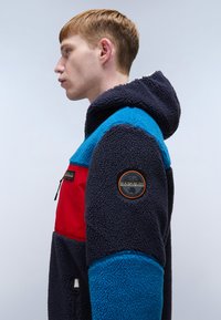 Fleece jacket with a navy body, blue accents, and a red chest pocket. Features a hood and a logo patch on the left sleeve.