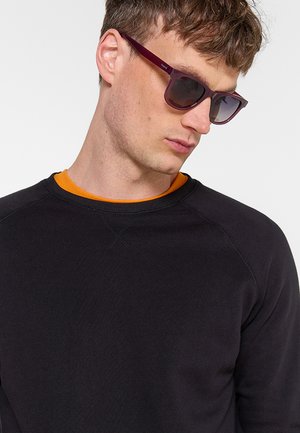 Uomo che indossa occhiali da sole viola scuro, maglione nero con scollo a giro e camicia arancione sotto, guarda in basso verso sinistra.