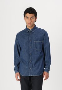 Camisa de denim en azul oscuro, con cierre de botones en el frente, cuello apuntado, un bolsillo en el pecho y puños con botones y detalles de costura visibles.