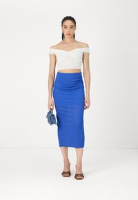 Balta off-the-shoulder īsā topa ar volānu malām, apvienots ar pieguļošu, līdz ceļiem garu zilu svārku un brūnām sandālēm ar siksniņām, tur rokās zilu clutch.