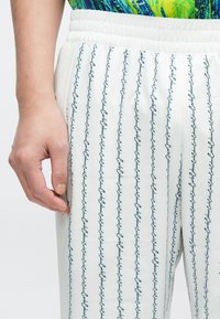 Valged püksid, millel on vertikaalne sinise kursiivkirjaga muster ja kummiga vöökoht. Sile pind, lähivaade, kus käsi puudutab kangast.