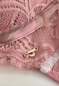 Close-up of pink lace fabric with mesh details and a small gold metal decorative charm attached near a stitched edge.