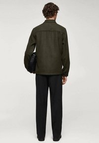 Olivengrøn uldjakke med krave, knappede manchetter og lige snit. Båret over sorte bukser, holdt med en sort håndtaske.