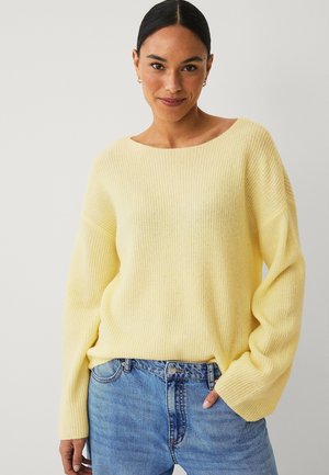 Kvinde med mørkt hår iført en lys gul strikket sweater og blå jeans, stående med den ene hånd i lommen og med et blidt smil mod en ensfarvet baggrund.