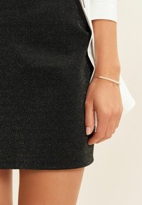 Zwarte glinsterende minirok, slim fit, gladde textuur. Draag met een delicate zilveren armband met kleine witte kralen en een hartaccent.
