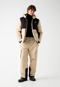 Beige en zwarte pufferjack met een capuchon, voorzien van ritsen en een logo, gecombineerd met beige cargobroek en zwarte laarzen. Soepele textuur.