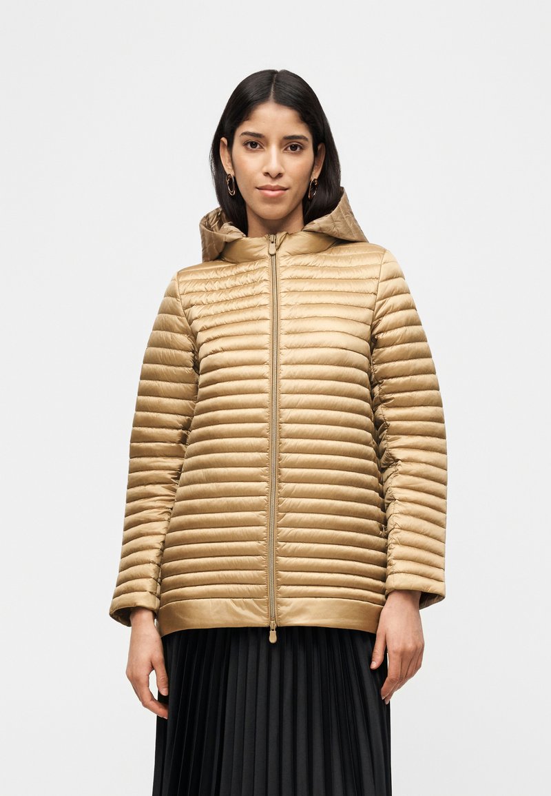 Γυναίκα που φοράει χρυσό καπιτονέ μπουφάν με κουκούλα και φερμουάρ, συνδυασμένο με μαύρη πλισέ φούστα, στέκεται μπροστά σε απλό λευκό φόντο.