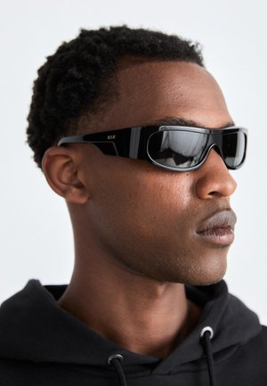 Lunettes de soleil noires au design sleek et angulaire. Cadre brillant et verres teintés foncés. Petit logo doré RSF sur le côté.