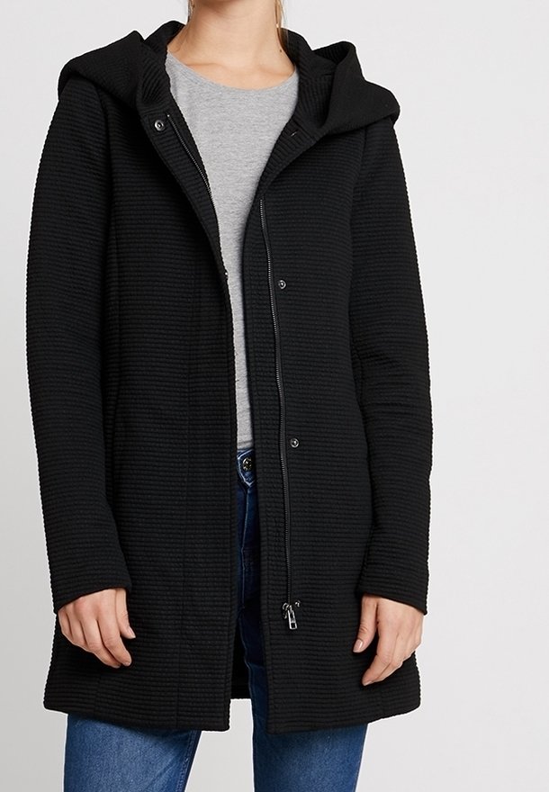 Cappotto nero con texture e cappuccio, dotato di una chiusura lampo anteriore e bottoni a pressione. Indossato sopra un top grigio e jeans blu.
