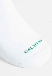 Calcetín blanco de algodón con textura acanalada y un logotipo verde que dice "CALSTAY" cerca del área de los dedos. Longitud estándar de caña.