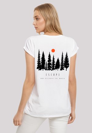 Frau mit blonden Haaren, die ein weißes T-Shirt mit schwarzen Kiefern, einer orangefarbenen Sonne und dem Text "ESCAPE" sowie "NOW DISCOVER THE WORLD." trägt.