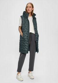 Gepolsterte ärmellose Weste in dunkelgrün mit hohem Kragen, zwei seitlichen Taschen und gestepptem Material. Kombiniert mit grauen Hosen und weißen Turnschuhen.