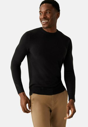 Black, long-sleeve sweater with a round neckline, made of a smooth, fitted fabric. Paired with light beige pants. Casual design.
